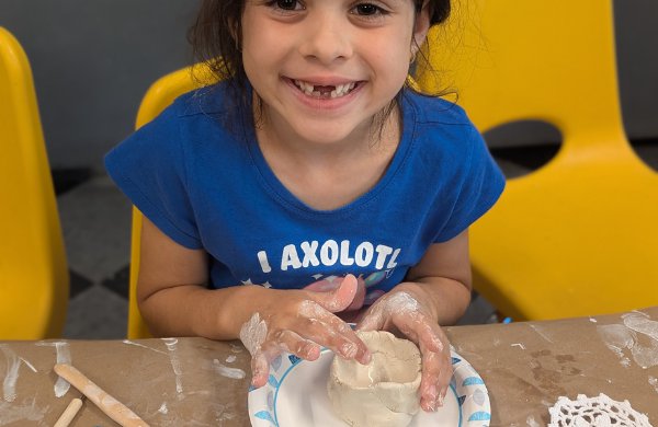 A child sitting at a table, looking at the camera smiling while making a clay pot. 