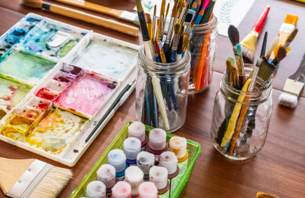 Art materials spread out on a table, such as brushes, paint and colored pencils. 