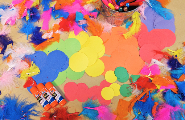 A variety of colorful feathers, paper circles and glue sticks scattered across a table with a bucket of markers. 
