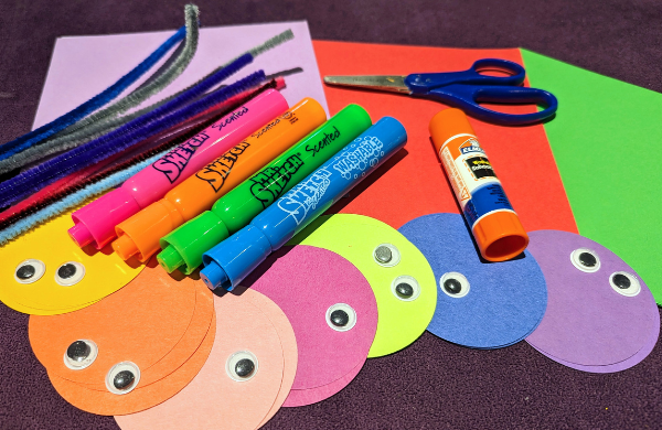 A pile of colorful construction paper sheets and circle, pipecleaners, markers, googly eyes, a glue stick and a scissor. 