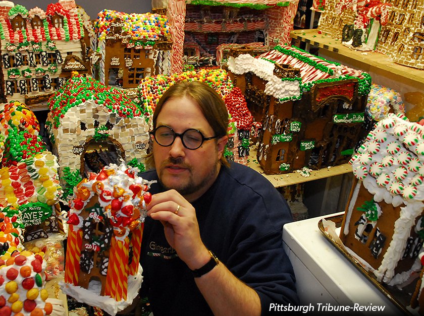 Man surrounded by decorated gingerbread houses while he decorates a gingerbread house. 