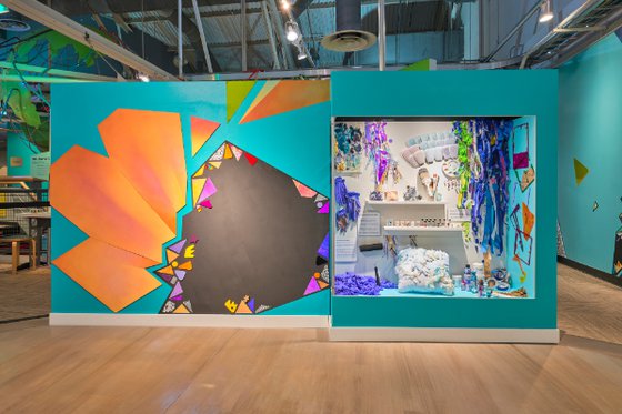 Two blue walls, one featuring geometric shaped artwork and a plexiglass display featuring ribbon and art tools. 