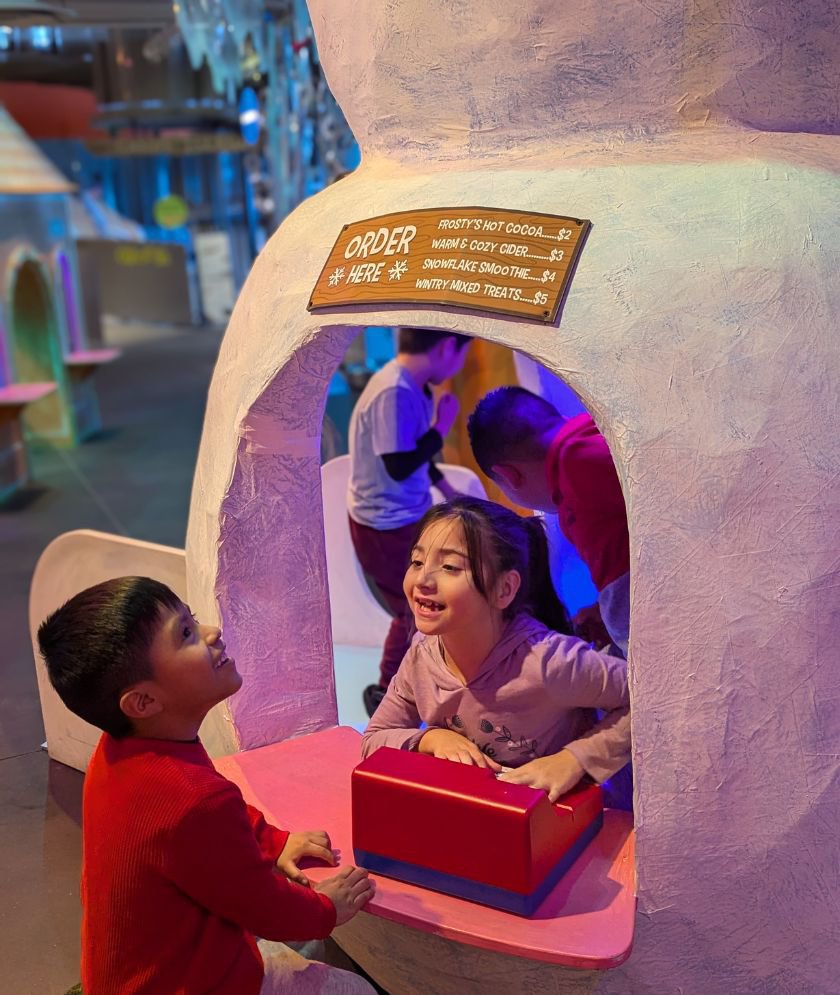 A child inside a cafe shaped as a snowman standing behind a register looking at another child on the outide. 