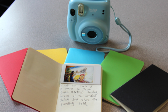 Six leather notebooks, one open with a picture of a painting in front of a polaroid camera. 