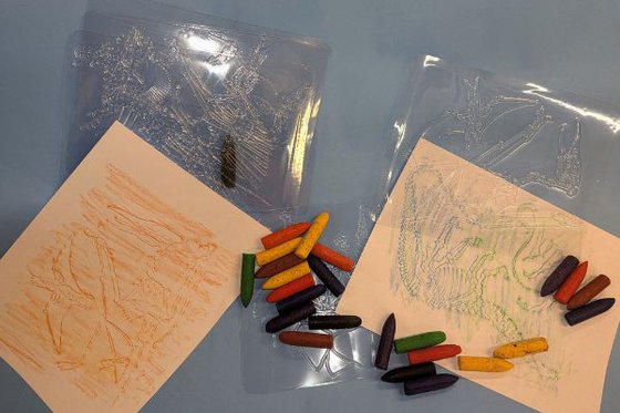 Clear plastic sheets with raised dinosaur designs for crayon rubbings, with crayons and paper.
