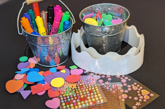 Two silver buckets filled with markers, foam stickers, gems, and two white paper crowns. 