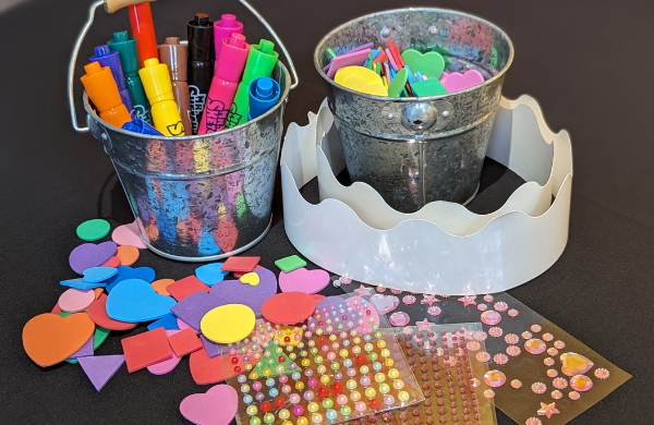 Two silver buckets filled with markers, foam stickers, gems, and two white paper crowns. 