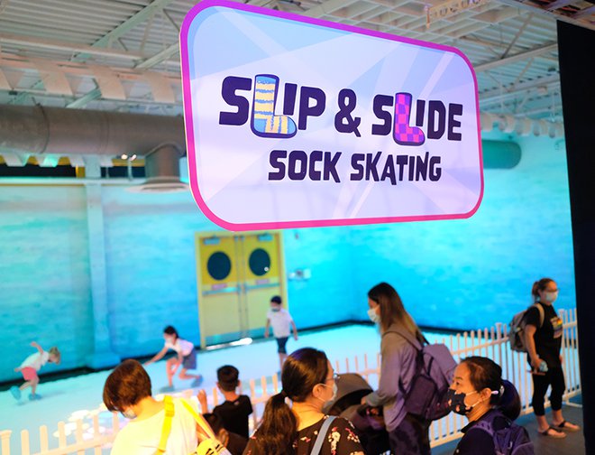 Entrance to exhibit "Sock Skating" featuring multiple adults watching children slide on the white floor while wearing face masks. 