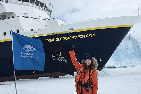 Woman in red parka standing in front of National Geographic Explorer ship alongside blue Lindblad Expeditions banner.
