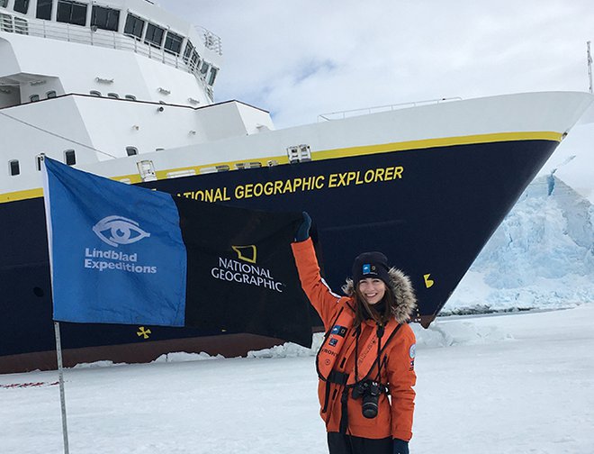 Woman in red parka standing in front of National Geographic Explorer ship alongside blue Lindblad Expeditions banner.