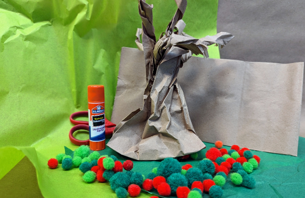 A bare tree made from brown paper surrounded by red and green pom poms, a scissor and a glue stick. 