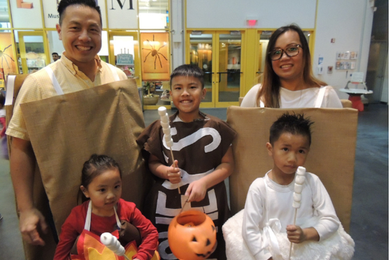 Two adults, at LICM, smiling and dressed as graham crackers standing behind three children dressed as a Hershey Chocolate bar, a bonfire and a marshmallow. 