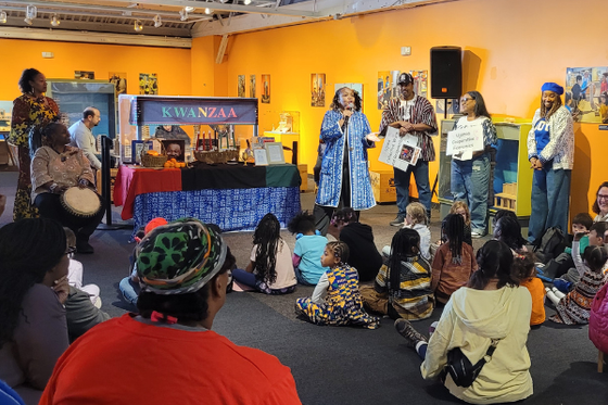 A group of children and adults sitting on a carpet looking at a group of speakers for a Kwanza celebration. 