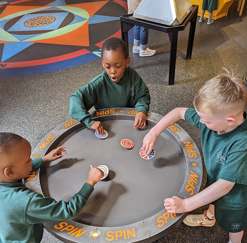 Three children standing at a circular table spinning tops. 