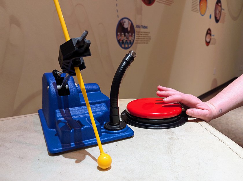 An adaptive tool used with a clamp holding a drum stick and a large button being pressed by a hand. 