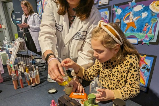 An adults dresssed in a lab coat standing behind a table next to a child helping them mix components for make up. 