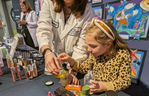 An adults dresssed in a lab coat standing behind a table next to a child helping them mix components for make up. 