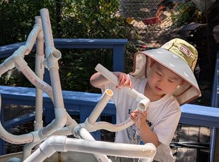 Young child focused on connecting plastic pipes on a water table, while water splashes around them. 