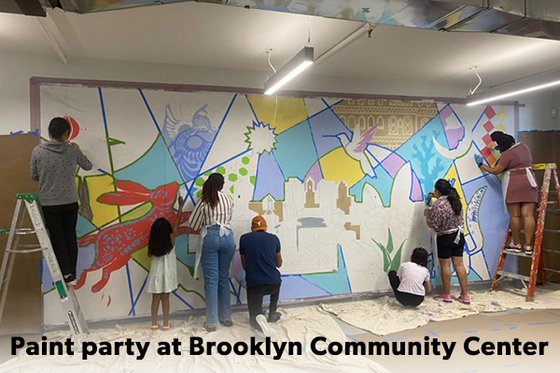 A group of seven people, two standing on a ladder, painting a mural of a landscape and colorful animals with the text Paint party at Brooklyn Community Services.