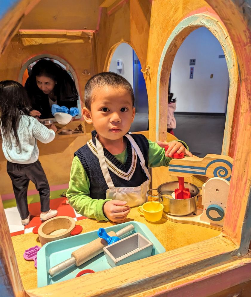 A child inside a bakery shaped as a gingerbread house looking at the camera while wearing a white apron and using a stand mixer. 