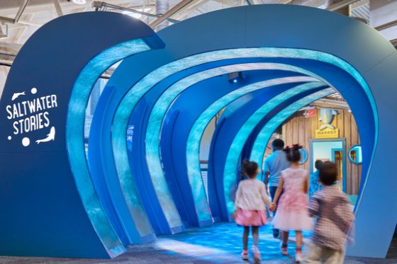 A large rib-like hollow blue structure made to mimic waves with text “Saltwater Stories” and two fish on the left side with a group of people walking underneath the structure.