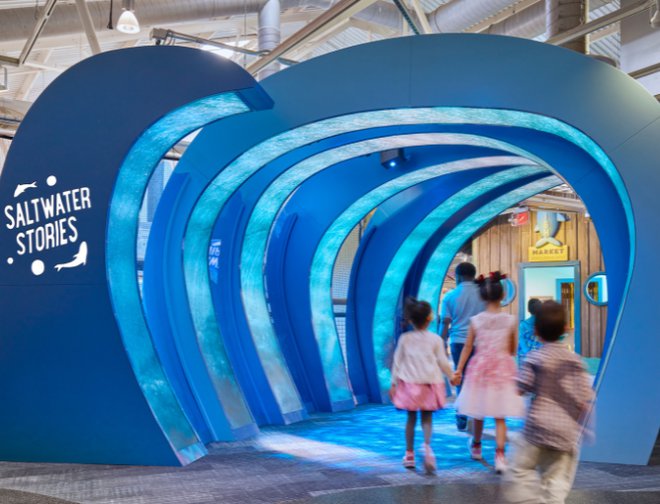 A large rib-like hollow blue structure made to mimic waves with text “Saltwater Stories” and two fish on the left side with a group of people walking underneath the structure.