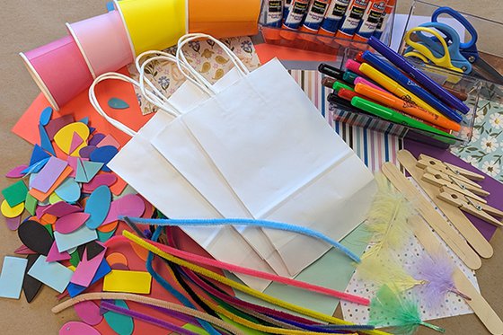Stack of white bags surrounded by craft items including markers, glue sticks, stickers and ribbons.