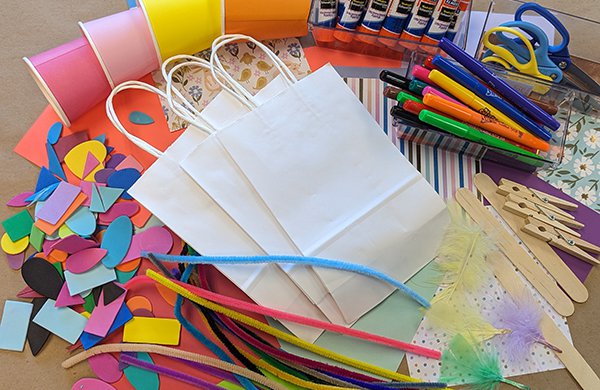 Stack of white bags surrounded by craft items including markers, glue sticks, stickers and ribbons.