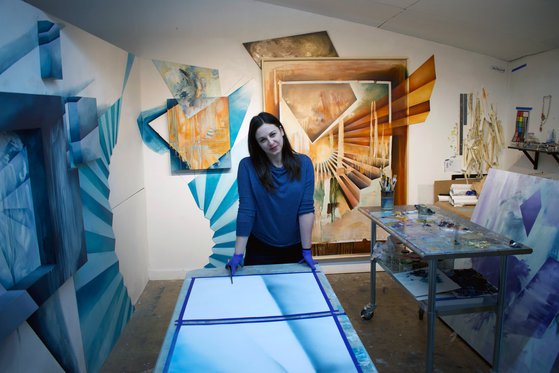 Kelly Olshan stands in her studio behind a table. the room is filled with abstract artwork.