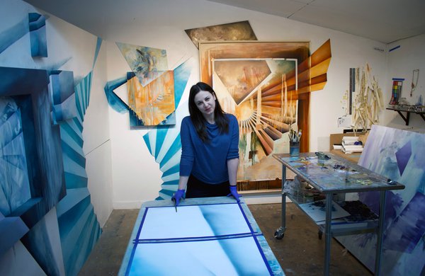 Kelly Olshan stands in her studio behind a table. the room is filled with abstract artwork.