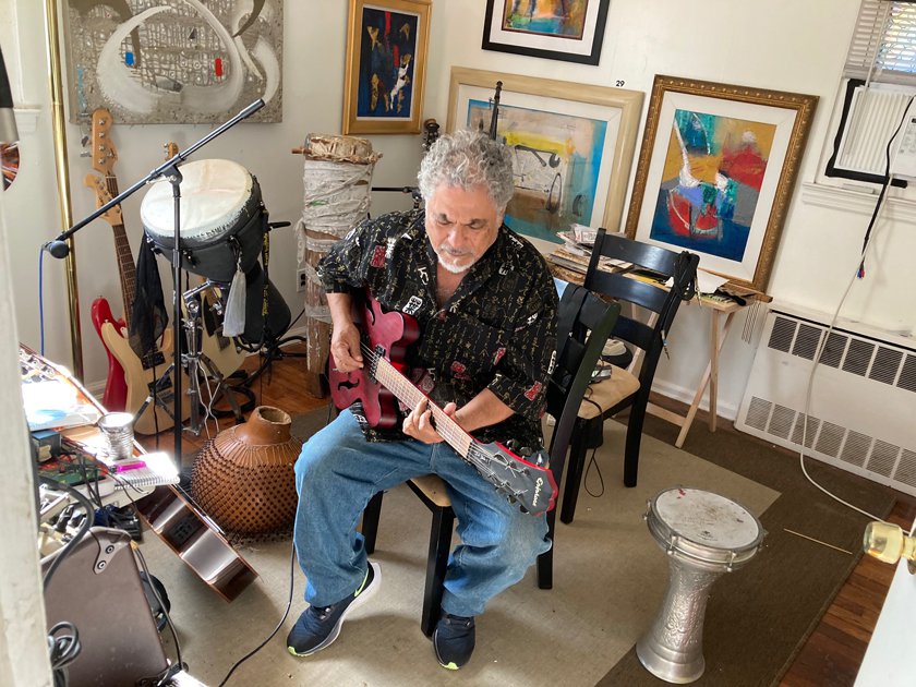 Man playing guitar in room surrounded by variety of drums and guitars.  Artwork is on all walls.