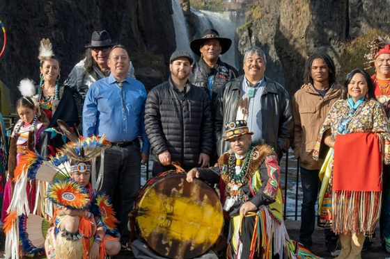A group of people standing in front of a waterfall, some wearing typical western clothes and some wearing traditional indigenous clothing. 