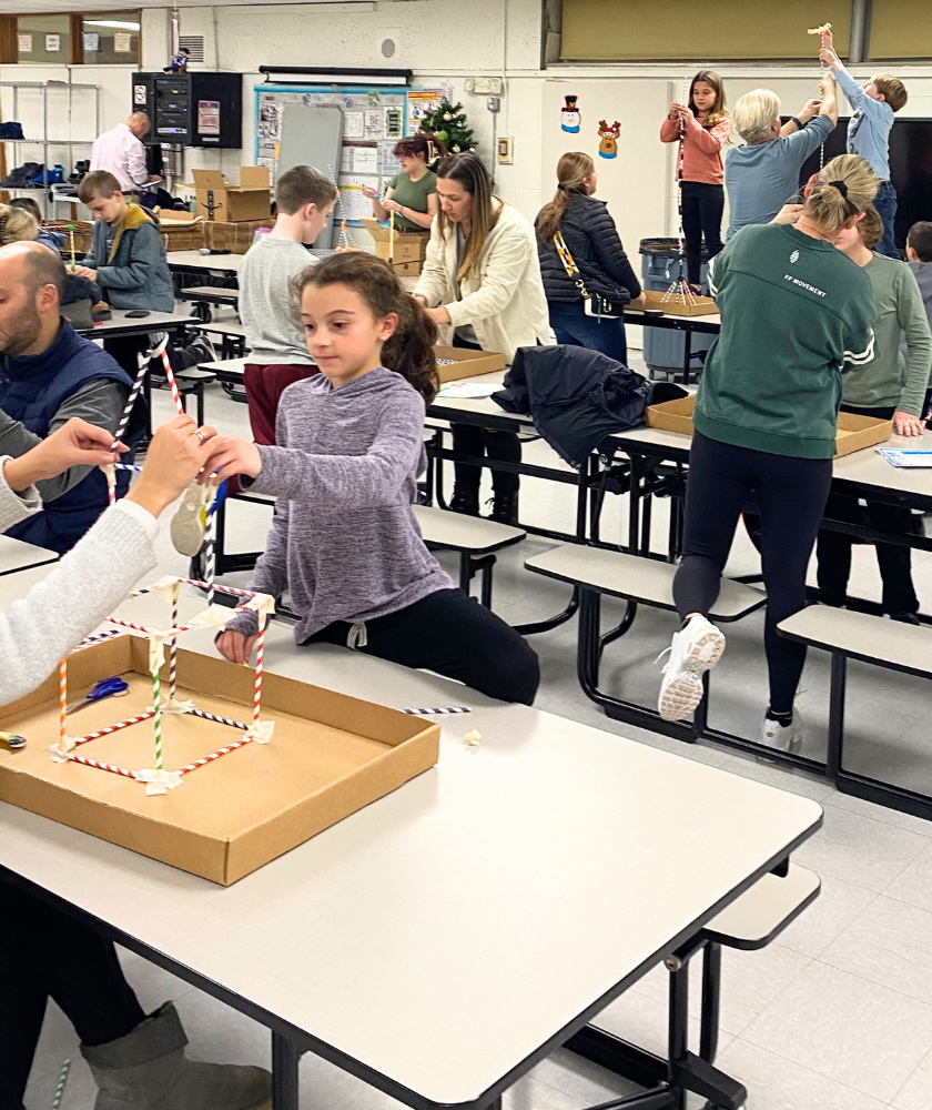 Children and adults standing and sitting at large tables building structures from paper straws. 