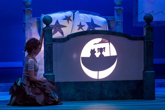 Actress on stage kneeling bedside a bed with a shadow design projected on footboard.