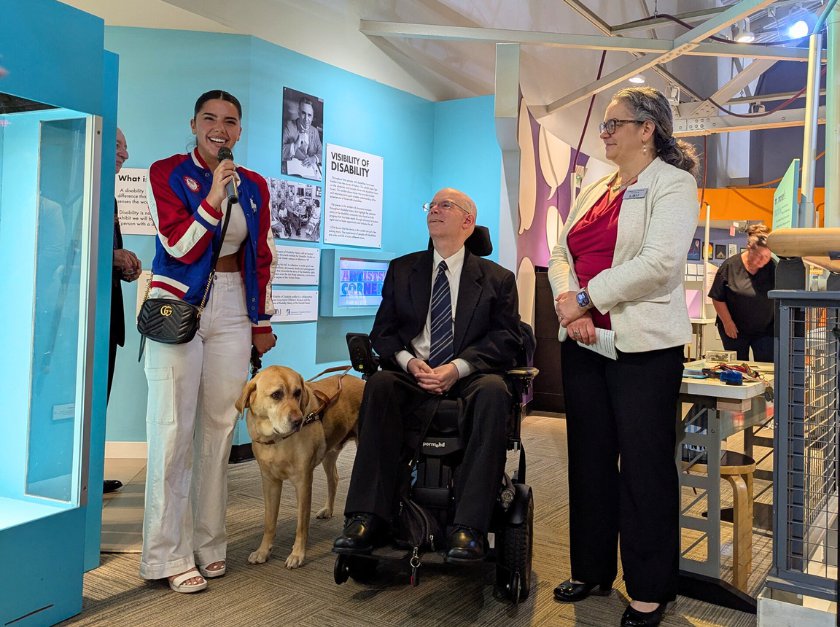 A woman holding a mic with a service dog, a man who uses a wheelchair, and a woman side by side in the Visability of Disability exhibit. 