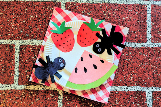 A red and white plaid patterned paper with paper slices of watermelon, strawberries, and ants. 