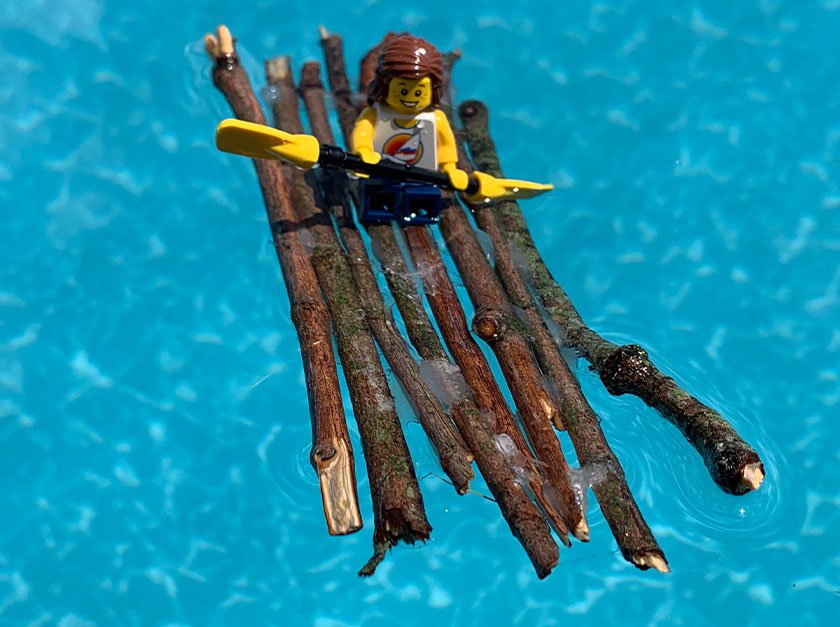 A raft made from sticks with a plastic person sitting on top with an oar while floating on water. 