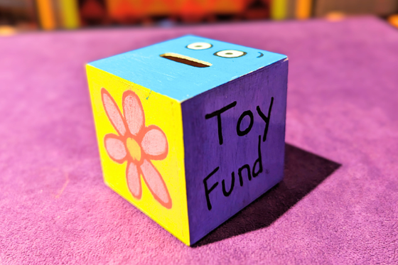 A cube box with a small open slot at the top and each side of the cube decorated and the text "Toy Fund." 
