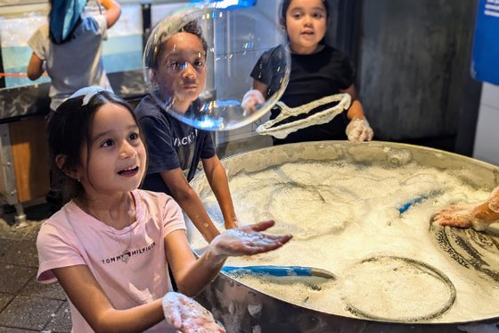 Three children look at a floating bubble with foam table in background. 