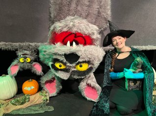 A small and large puppet of a bat upside down next a person in a witch costume. 