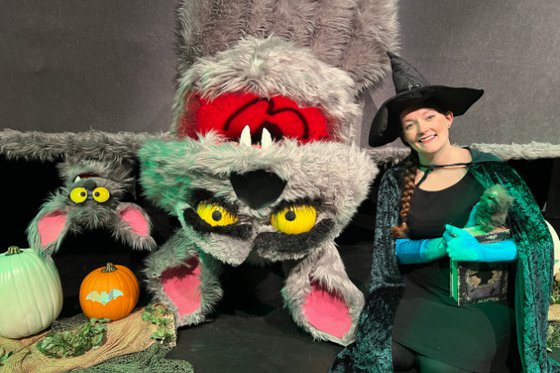 A small and large puppet of a bat upside down next a person in a witch costume. 