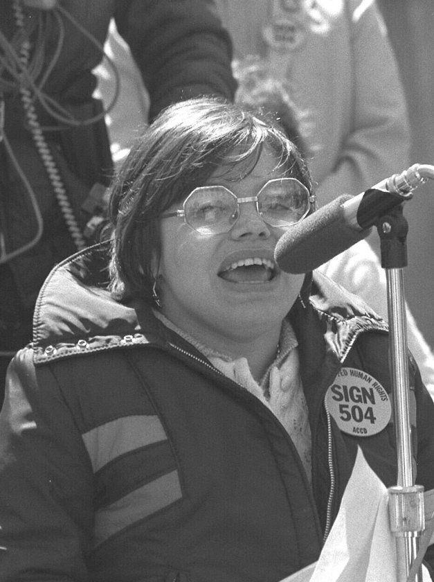 Black and white photo of Judy Heumann speaking into a microphone. 