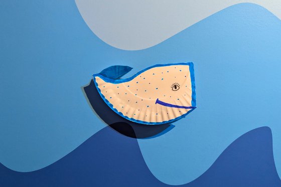 White paper plate folded into a whale shape, colored with blue markers. Set on blue gradient wall with wave design.