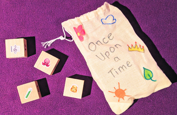 A small decorated cloth bag with text "Once Upon a Time",  and four wooden cubes with a picture of a musical note, octopus, test tube, and Queen.