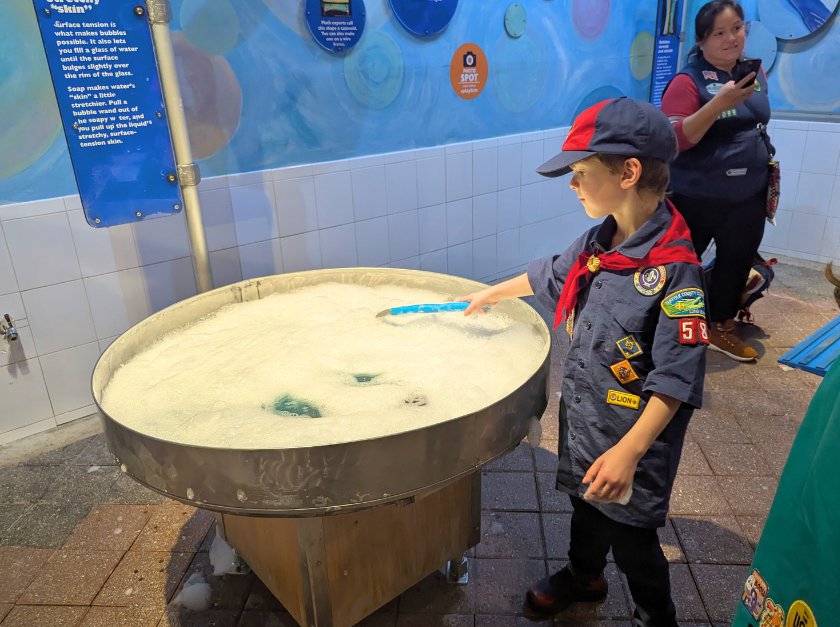 A boy scout wearing a dark blue uniform using a bubble wand at a bubble table. 