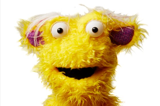 A fuzzy yellow puppet troll with his mouth open. Gruff has pink ears, big white eyes, and white hair on his head. 