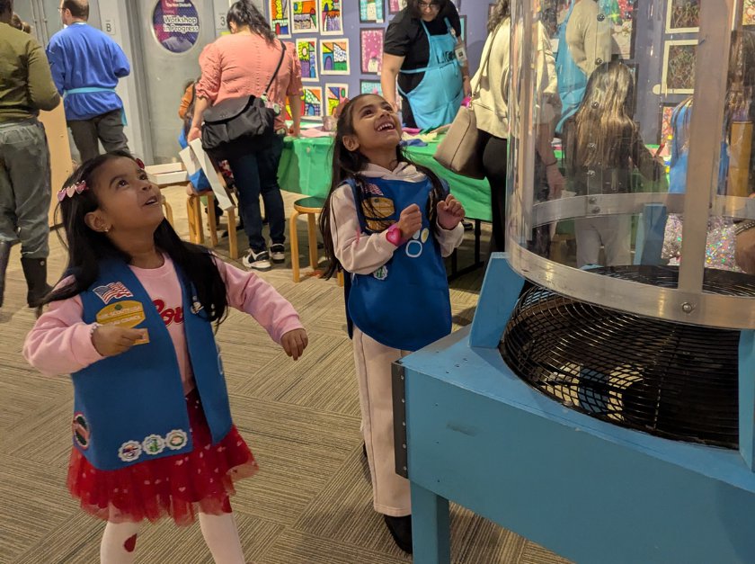 Two girls wearing blue vests looking up at a wind tunnel. 