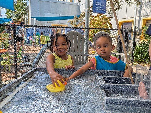 Two children looking at the camera while standing at a water table outside and holding onto a rubber duck. 