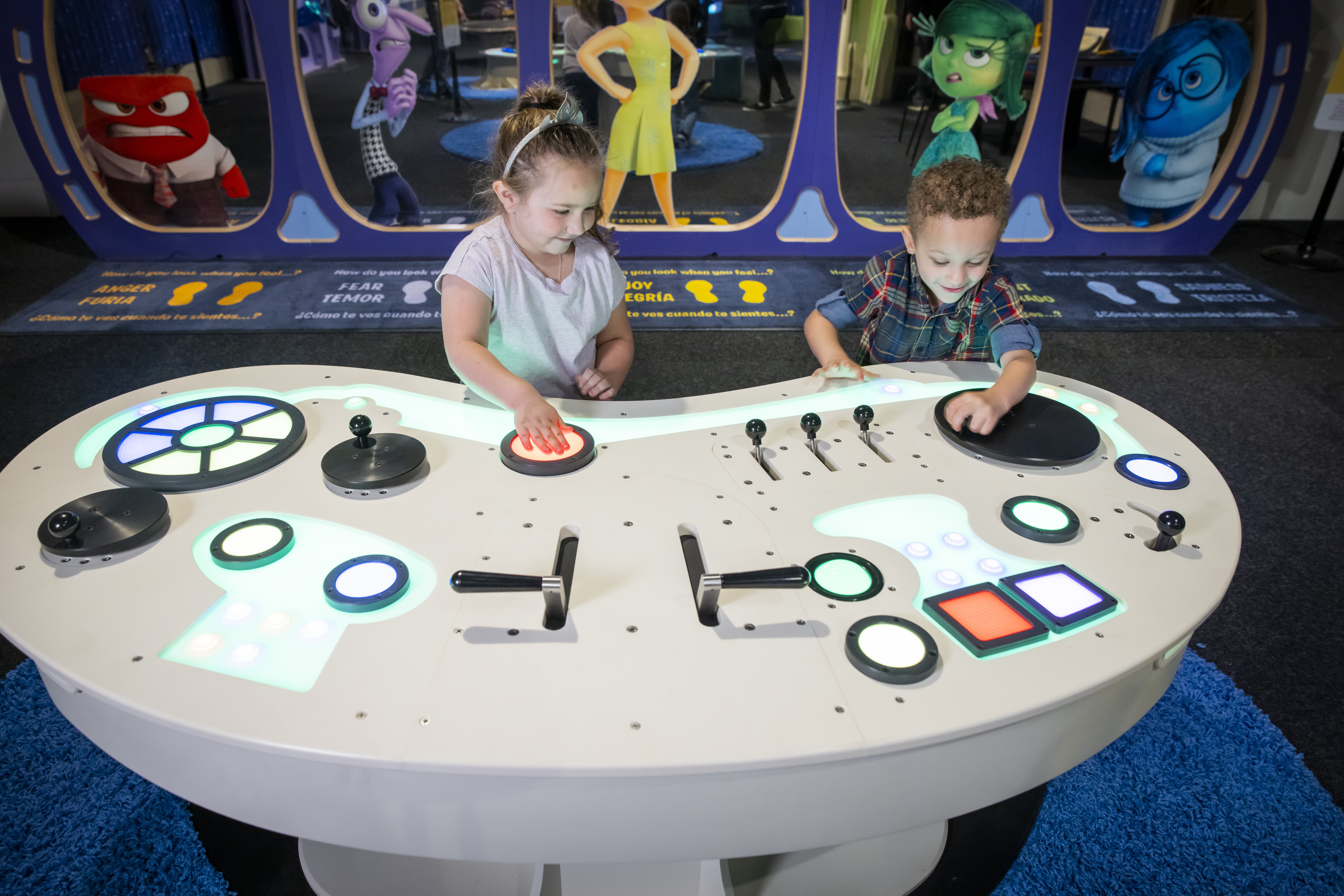 Two children play at a glowing, interactive control table, with large characters from the movie Inside Out on screens behind them.