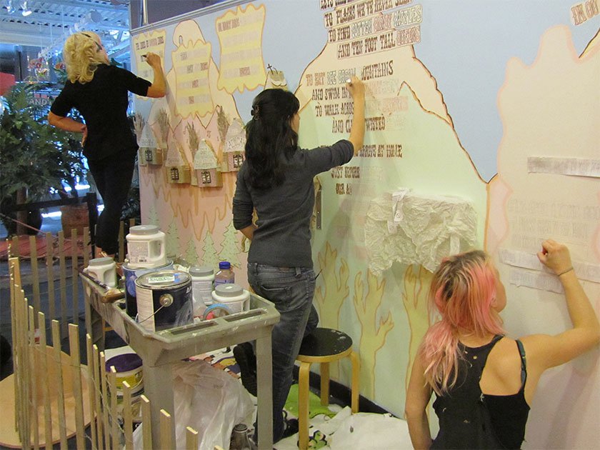 Three artists creating a mural featuring homes, trees, mountains and quotes using markers. 